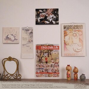 Op de afbeelding: Vier wandkalenders met vintage illustraties, waaronder een kalender met katten, een Japanse prent, een poster over het dagelijks leven in de jaren 60 en een Alphonse Mucha Salon des Cent kalender, naast een gouden lijst en twee kokeshi-poppen.