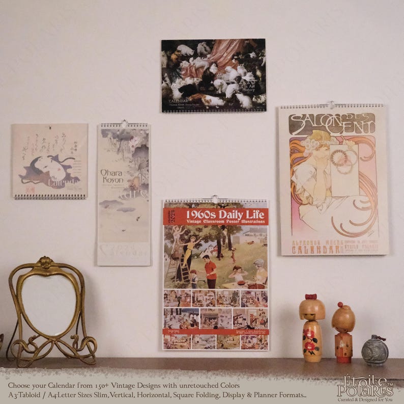 Op de afbeelding: Vier wandkalenders met vintage illustraties, waaronder een kalender met katten, een Japanse prent, een poster over het dagelijks leven in de jaren 60 en een Alphonse Mucha Salon des Cent kalender, naast een gouden lijst en twee kokeshi-poppen.