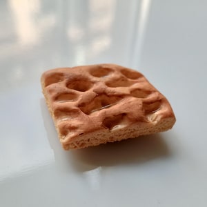 May include: A square, light brown pastry with a textured surface and a pattern of rounded indentations. The pastry is on a light grey surface.