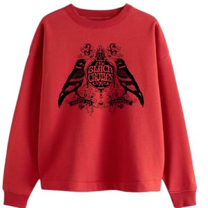 May include: A red sweatshirt featuring a black graphic of two crows flanking a circular emblem with the text "The Black Crowes" and the words "Hard to Handle". The sweatshirt has a crew neck and long sleeves.