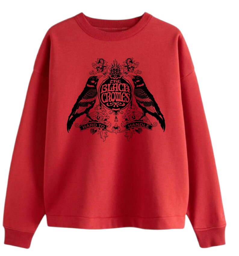 May include: A red sweatshirt featuring a black graphic of two crows flanking a circular emblem with the text "The Black Crowes" and the words "Hard to Handle". The sweatshirt has a crew neck and long sleeves.