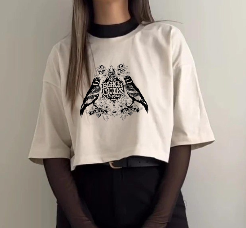 May include: Cream-colored, short-sleeved t-shirt with a black graphic featuring two crows flanking a crest that reads "The Black Crows." The text "Hard to Handle" is below the crows. The shirt has a relaxed fit.