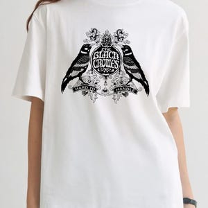 May include: White t-shirt featuring a black graphic design. The design includes two crows flanking a circular emblem with the text "THE BLACK CROWES" and the words "HARD TO HANDLE" below. The shirt has short sleeves.