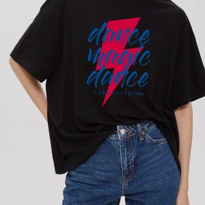 Pode incluir: Uma camiseta preta oversized com um gráfico de raio vermelho e o texto "dance magic dance" em azul. O texto "LABYRINTH 1986" é impresso abaixo do gráfico.