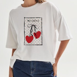 Puede incluir: Camiseta blanca con un estampado gráfico de dos corazones rojos con una cinta negra y el texto "Ma Chérie" en un rectángulo negro.