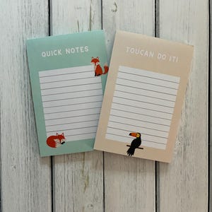 May include: Two notepads with lined pages. One is mint green with the text "QUICK NOTES" and a fox illustration. The other is beige with the text "TOUCAN DO IT!" and a toucan illustration. Both are on a white wooden surface.