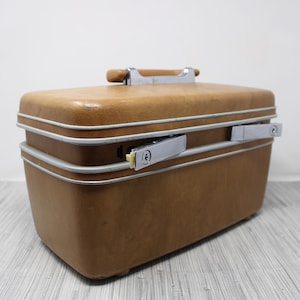 May include: A vintage brown suitcase with a silver trim and two silver latches. The suitcase has a wooden handle and is rectangular in shape. The suitcase is made of a durable material and appears to be in good condition.