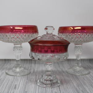 May include: Three glass candy dishes with a red and clear design. The dishes have a pedestal base and a round bowl. The center dish has a lid.