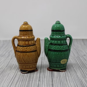 May include: Two ceramic salt and pepper shakers shaped like teapots. The shaker on the left is brown with black and white details. The shaker on the right is green with black and white details. Both shakers have a sticker on the bottom.