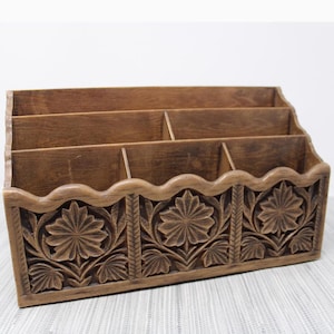 May include: A wooden desk organiser with four compartments. The front features carved floral designs. The organiser is made of a medium-toned wood and has a scalloped edge along the top of the floral carving.