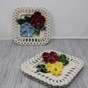May include: Two white ceramic square plates with a decorative lace border. Each plate features a 3D floral design with red, yellow, blue, and green flowers.