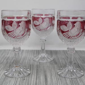 May include: Three clear crystal wine glasses with a ruby red interior. Each glass features an etched design of a bird perched on a branch with leaves. The glasses have a detailed stem and base.