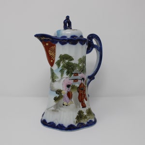 May include: A white porcelain teapot with blue and gold trim. The teapot features a hand-painted scene of Japanese women in traditional clothing walking through a landscape with trees and a pagoda.