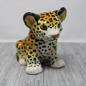 May include: A ceramic jaguar figurine with a spotted pattern in yellow, black, and white. The figurine is in a seated position with its head turned slightly to the side, showcasing its detailed facial features and large ears.