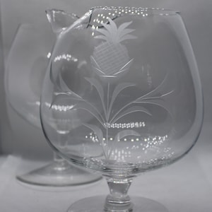May include: Two clear glass brandy snifters with etched pineapple designs. The snifters have a round bowl and a tall stem.