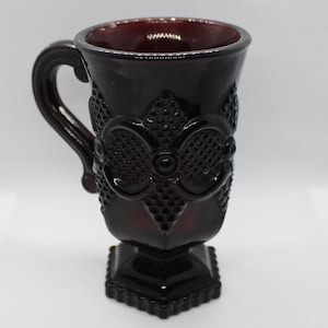 May include: A dark red glass mug with a footed base and a handle. The mug has a decorative pattern of raised diamonds and a floral design.