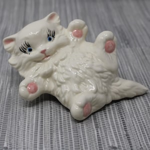 May include: A white ceramic cat figurine lying on its back with its paws in the air. The cat has blue eyes, pink nose and paw pads, and black eyelashes. The figurine is a decorative item.