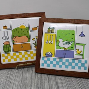 May include: Two framed ceramic tiles with a kitchen theme. One tile features a pig and a pumpkin, the other a duck and pineapples. Both have a wooden frame and a colorful, cartoon-like design.