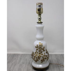 Peut inclure: Une lampe de table en verre blanc avec un motif de feuilles dorées. La lampe a une base de couleur laiton et une douille standard. La lampe mesure environ 38 cm de haut.