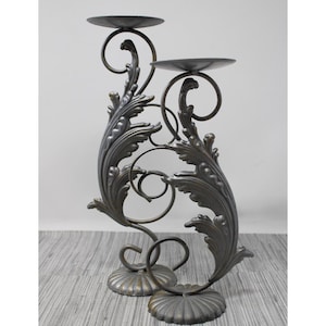 May include: A decorative, two-tiered metal candle holder. The candle holder features ornate, leaf-like designs and swirling accents. The base and candle platforms are circular. The color is a muted bronze.