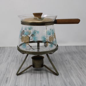 May include: A glass teapot with a wooden handle and a pine cone design. The teapot is on a metal stand with a candle holder.