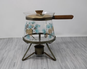 May include: A glass teapot with a wooden handle and a pine cone design. The teapot is on a metal stand with a candle holder.