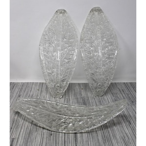 May include: Three clear glass leaf-shaped decorative pieces. Two are upright, and one is horizontal. Each piece has a textured, etched design resembling veins and patterns of a leaf. The pieces are on a striped surface.