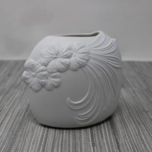 May include: A white ceramic vase with a rounded shape and a rectangular opening. The vase features a floral design with three embossed flowers and flowing, textured lines. The vase is approximately 6 inches tall.