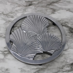 May include: A silver-colored trivet featuring three detailed seashell designs within a circular frame. The trivet is made of metal and is designed to protect surfaces from hot items. The background is a marble-patterned surface.