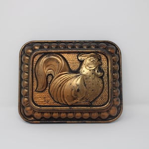 May include: A gold-colored metal rooster figurine with a detailed design. The rooster is framed by a decorative border of small beads.