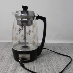 May include: Vintage electric coffee percolator with a clear glass carafe decorated with gold and black starbursts. The percolator has a black base and handle, a silver lid, and a black power cord. A classic kitchen appliance.