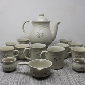 May include: A complete vintage stoneware tea set in a soft, creamy white. The set includes a teapot, cups, mugs, and a small creamer. Each piece is adorned with a delicate floral pattern, perfect for a classic kitchen.