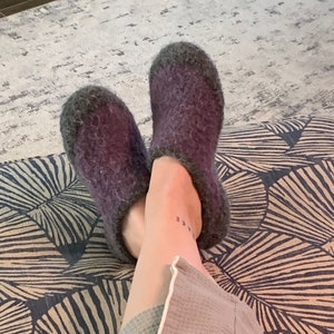 May include: A pair of purple felted wool slippers with gray trim. The slippers are shown on a patterned surface, suggesting a cozy indoor setting. The slippers appear soft and comfortable, ideal for relaxation.