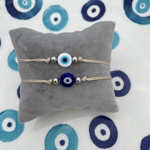 May include: Two evil eye charm bracelets. Each bracelet has a beige cord, silver beads, and a glass evil eye charm. One charm is white and light blue, the other is dark blue. The bracelets are displayed on a grey cushion.