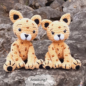 Op de afbeelding: Twee gehaakte luipaard amigurumi speelgoedjes die op een rots zitten. De speelgoedjes zijn beige met zwarte vlekken en hebben zwarte ogen en neuzen. De speelgoedjes zitten op een rots met een witte wolk met de tekst "Anleitung/Pattern" en de tekst "HäkelWorld" onder de wolk.