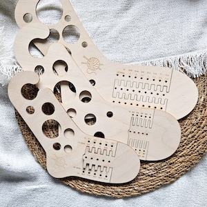 Könnte beinhalten: Drei Strickwerkzeuge aus Holz für Socken mit unterschiedlich großen Löchern und Messmarkierungen. Jedes Werkzeug hat einen gebogenen Griff und eine fußförmige Basis. Auf jedem Werkzeug ist eine Garn- und Nadelgrafik eingeätzt.