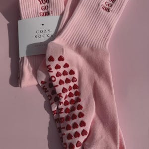 May include: A pair of light pink ribbed crew socks featuring the embroidered red text YOU GO GIRL on the cuff. The soles have red heart-shaped silicone grips and the text WOLVENKIND. A white paper label reads COZY SOCKS with a small heart icon.