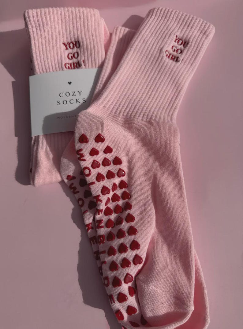 May include: A pair of light pink ribbed crew socks featuring the embroidered red text YOU GO GIRL on the cuff. The soles have red heart-shaped silicone grips and the text WOLVENKIND. A white paper label reads COZY SOCKS with a small heart icon.