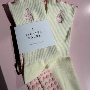 May include: A pair of cream-colored ribbed socks with pink lettuce-edge cuffs. The socks feature pink embroidered text reading GRL PWR with a heart above it. A white paper band is wrapped around the socks with the text PILATES SOCKS WOLKENBILDER.