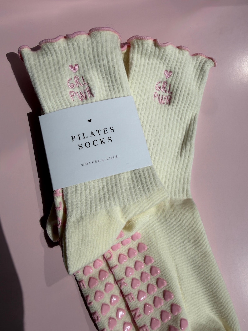 May include: A pair of cream-colored ribbed socks with pink lettuce-edge cuffs. The socks feature pink embroidered text reading GRL PWR with a heart above it. A white paper band is wrapped around the socks with the text PILATES SOCKS WOLKENBILDER.