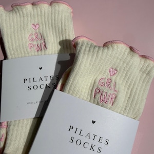 May include: A pair of cream-colored ribbed socks with pink lettuce-edge cuffs. Each sock features an embroidered pink heart and the text GRL PWR. The socks are packaged with a white paper band labeled PILATES SOCKS and WOLKENBILDER.