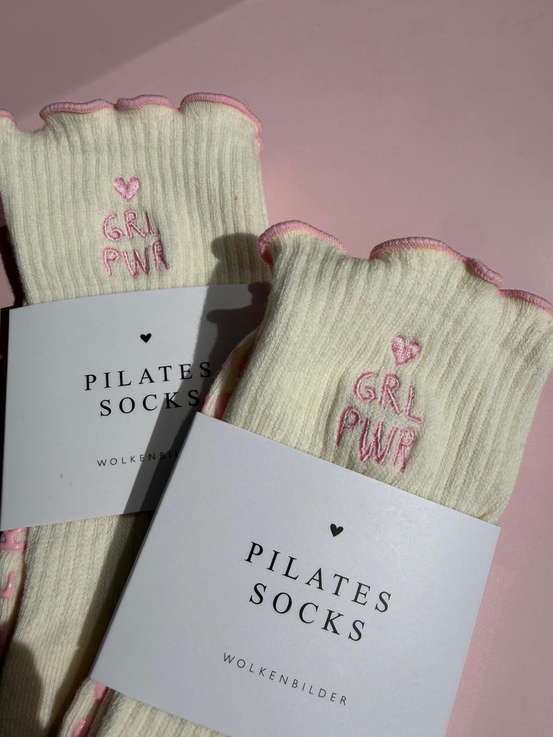 May include: A pair of cream-colored ribbed socks with pink lettuce-edge cuffs. Each sock features an embroidered pink heart and the text GRL PWR. The socks are packaged with a white paper band labeled PILATES SOCKS and WOLKENBILDER.