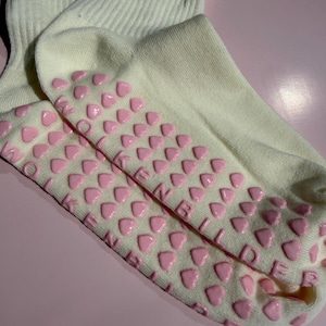 May include: Cream-colored ribbed socks featuring a pattern of small, raised pink heart-shaped silicone grips on the soles. The text WOLKENBILDER is embossed in pink lettering integrated among the heart grips on the bottom of the socks.