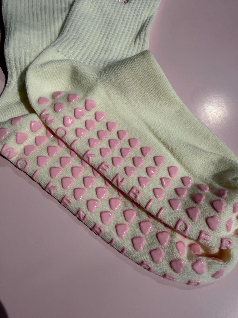 May include: Cream-colored ribbed socks featuring a pattern of small, raised pink heart-shaped silicone grips on the soles. The text WOLKENBILDER is embossed in pink lettering integrated among the heart grips on the bottom of the socks.