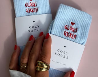 May include: A pair of light blue ribbed socks featuring red embroidered text that reads GLÜCKS SOCKEN with a small red heart above the words. Each sock is wrapped in a white paper band printed with the text COZY SOCKS and WOLKENBILDER.