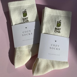 Könnte beinhalten: Zwei Paar cremefarbene gerippte Socken mit einem Matcha-Getränk-Motiv und dem Text "MAT CHA". Jedes Paar ist mit einem weißen Etikett mit der Aufschrift "COZY SOCKS" und "WOLKENBILDER" verpackt. Die Socken liegen auf einer rosa Oberfläche.