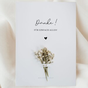 May include: A white card with the text "Danke!" and "F&Uuml;R EINFACH ALLES!" in black script. A small black heart is below the text. A small bouquet of white baby's breath flowers is below the heart. The text "WOLKENBILDER" is at the bottom of the card.