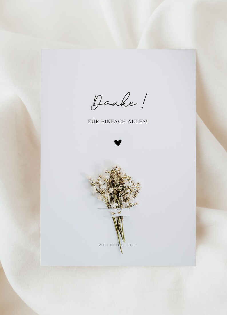 May include: A white card with the text "Danke!" and "F&Uuml;R EINFACH ALLES!" in black script. A small black heart is below the text. A small bouquet of white baby's breath flowers is below the heart. The text "WOLKENBILDER" is at the bottom of the card.