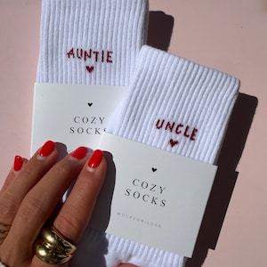 Könnte beinhalten: Zwei Paar weiße gerippte Socken, verpackt mit Papierbanderolen. Ein Paar zeigt den roten Sticktext AUNTIE mit einem kleinen Herz, das andere UNCLE mit einem Herz. Die Banderolen sind mit COZY SOCKS und WOLKENBILDER bedruckt.