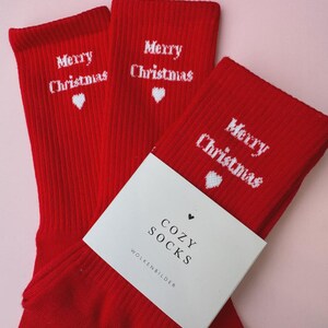 Könnte beinhalten: Drei Paar rote Weihnachtssocken mit dem Schriftzug "Merry Christmas" in Weiß und einem weißen Herz. Ein weißes Etikett trägt die Aufschrift "Cozy Socks" und "Wolkenbilder".